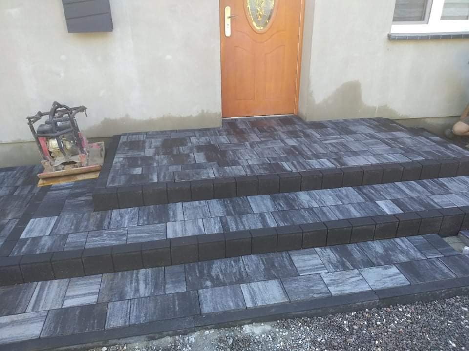 Schody wejściowe wyłożone szaro-czarną kostką brukową, z widoczną zagęszczarką w tle, przed drewnianymi drzwiami wejściowymi.