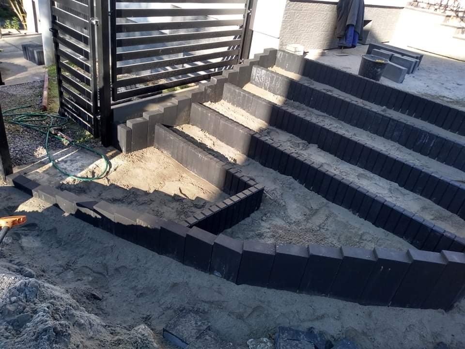Schody zewnętrzne w trakcie budowy, obłożone ciemnoszarą kostką obrzeżową, z wypełnieniem piaskiem, widoczna furtka ogrodzeniowa w tle.