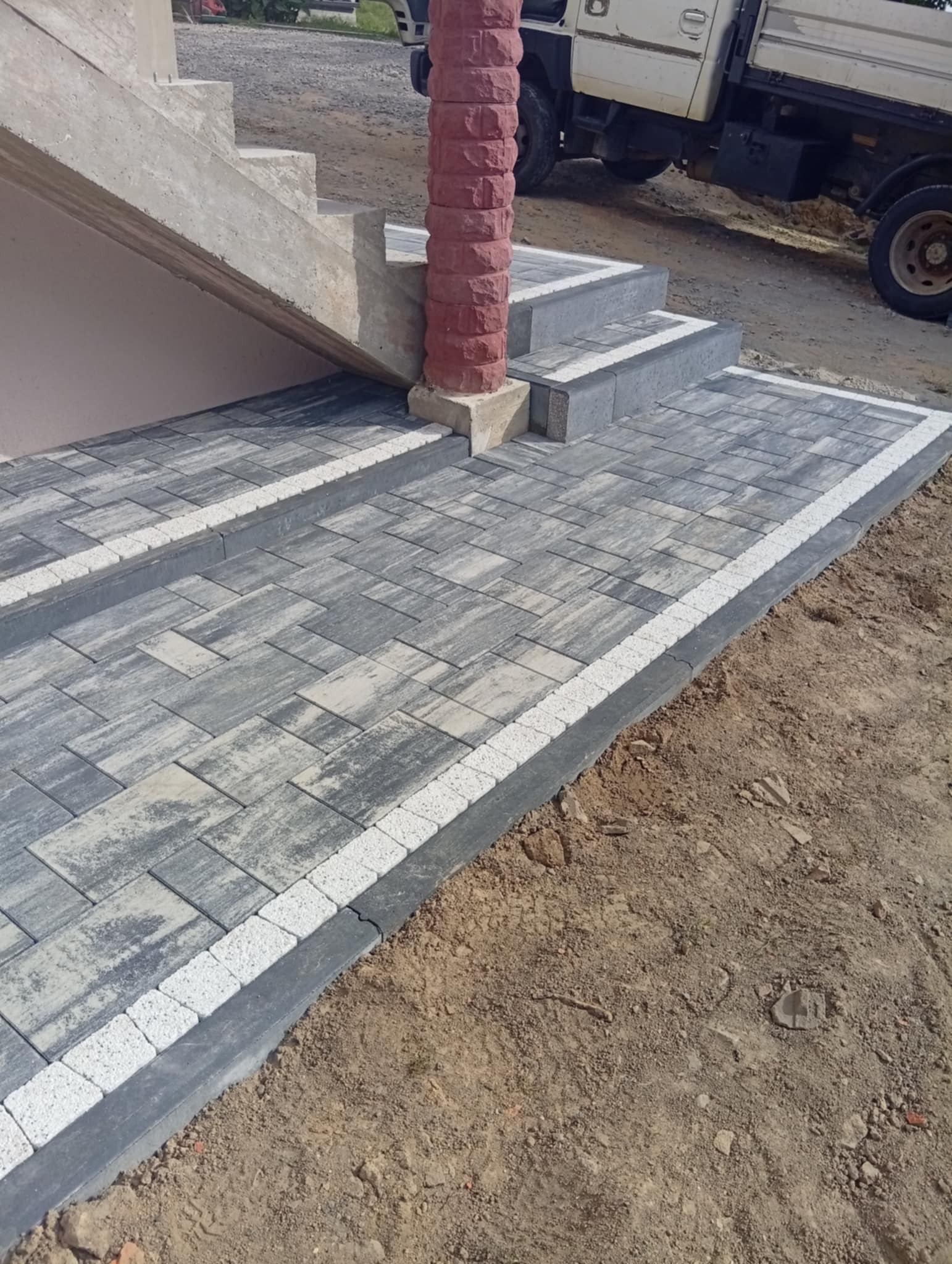 Ułożona kostka brukowa w szaro-białej kolorystyce prowadząca do betonowych schodów, z kolumną z czerwonej cegły. W tle zaparkowany biały samochód dostawczy.