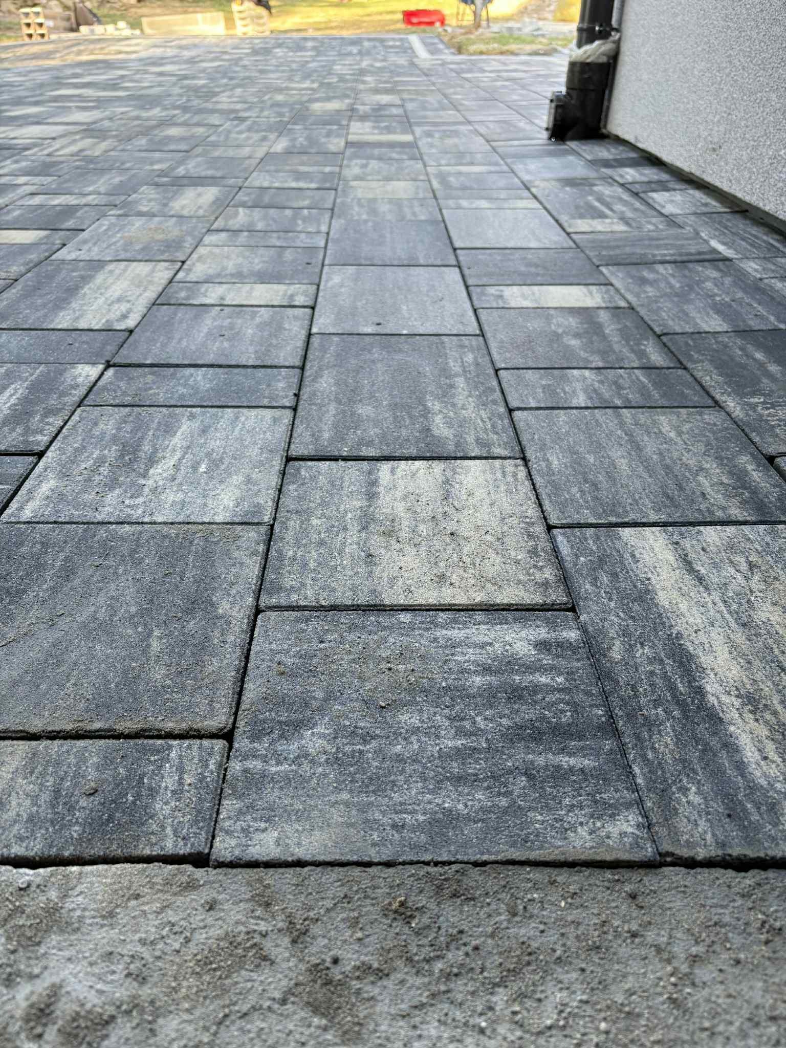 Ułożona kostka brukowa w odcieniach szarości, z widoczną fakturą kamienia. Układ prostokątny, perspektywa z dołu, z fragmentem ściany budynku w tle.