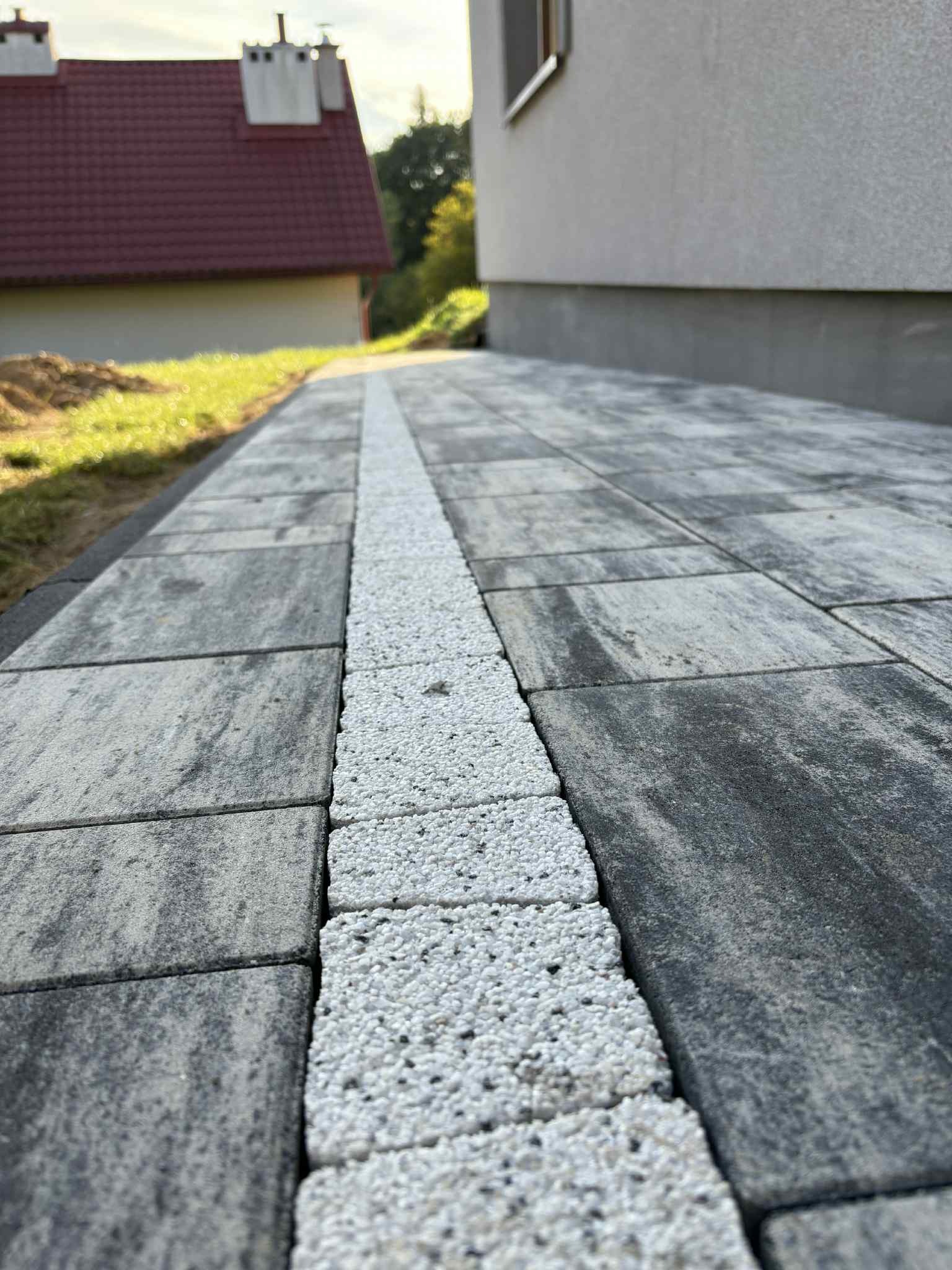 Ułożona kostka brukowa w odcieniach szarości z białym pasem, prowadząca wzdłuż ściany budynku i trawnika. Widok z niskiej perspektywy.