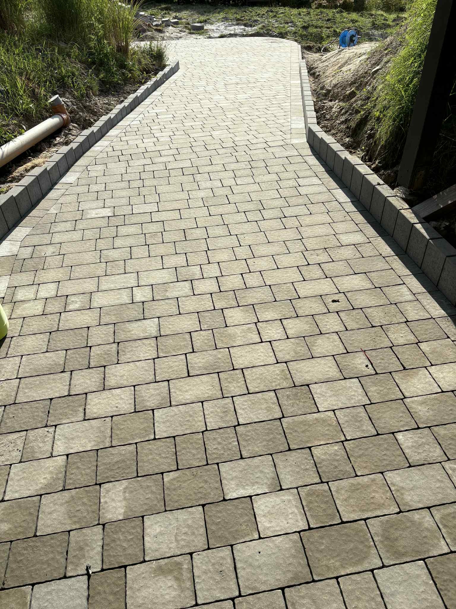 Ścieżka z jasnoszarej kostki brukowej, obrzeża z ciemniejszej kostki. Widok z perspektywy, trawa po bokach. Długa rura PVC leży przy ścieżce.