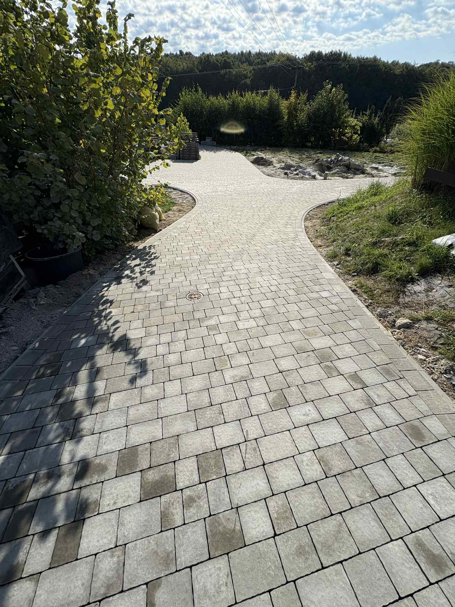 Ścieżka z kostki brukowej w ogrodzie, z widocznymi elementami zieleni i drzewami w tle. Kostka ułożona w regularny wzór, na rozwidleniu znajduje się studzienka.