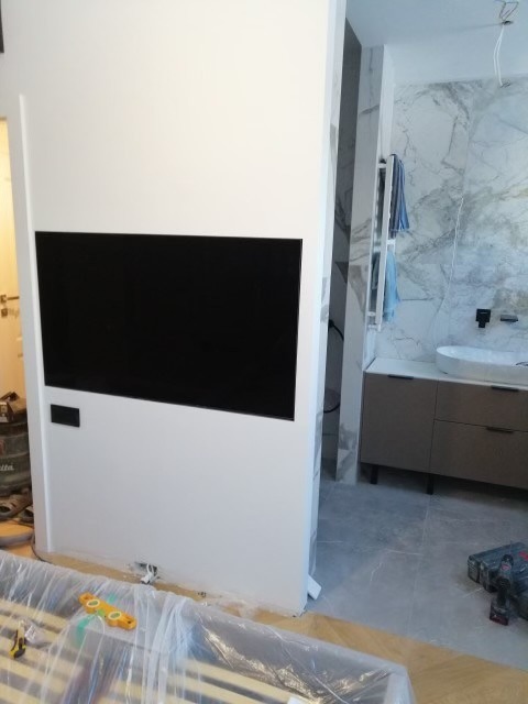 Biała ścianka działowa z wnęką na telewizor w trakcie montażu, widoczne narzędzia i elementy wykończeniowe, łazienka z marmurowymi płytkami w tle.