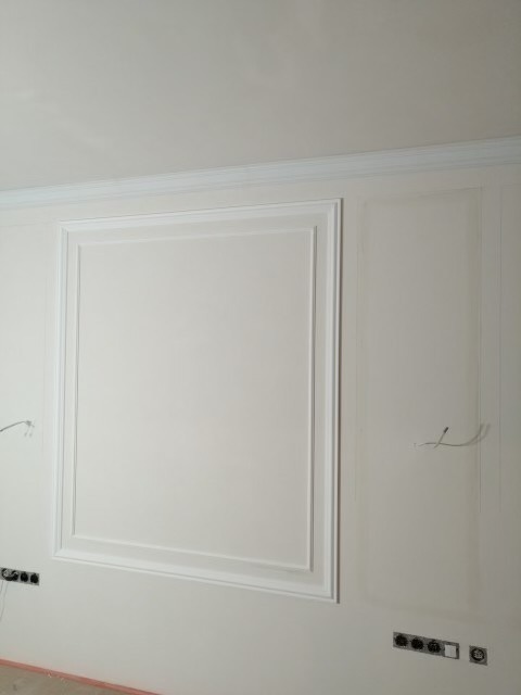 Ściana z dekoracyjnymi panelami w kolorze złamanej bieli, z widocznymi gniazdkami elektrycznymi i wystającymi kablami, ukazująca etap prac wykończeniowych.