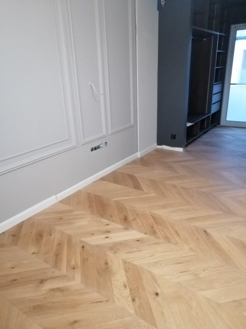 Parkiet ułożony w jodełkę w jasnym odcieniu drewna, szara ściana z dekoracyjnymi listwami i zabudowana szafa w tle.