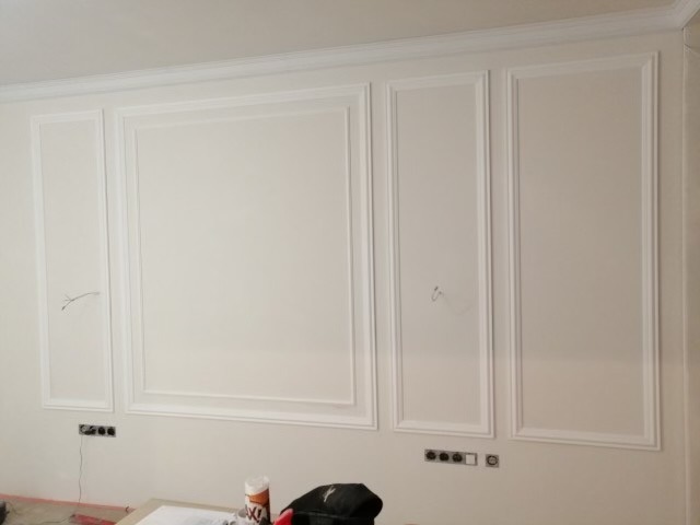 Ściana z zamontowanymi, białymi panelami dekoracyjnymi w stylu klasycznym, widoczne wystające przewody i gniazdka elektryczne, częściowo zasłonięte materiały budowlane na pierwszym planie.