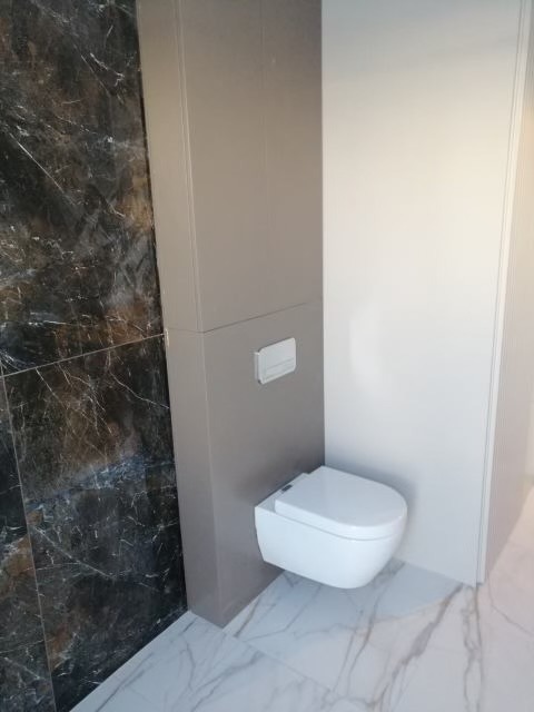 Nowoczesna toaleta podwieszana w łazience z marmurowymi płytkami na ścianie i podłodze, obok szafka w zabudowie.