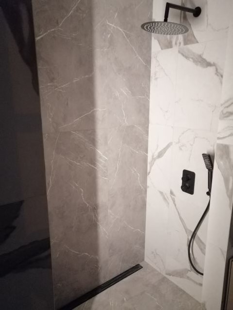 Wykończona kabina prysznicowa z szarymi i białymi płytkami imitującymi marmur, widoczny czarny odpływ liniowy i czarna armatura.