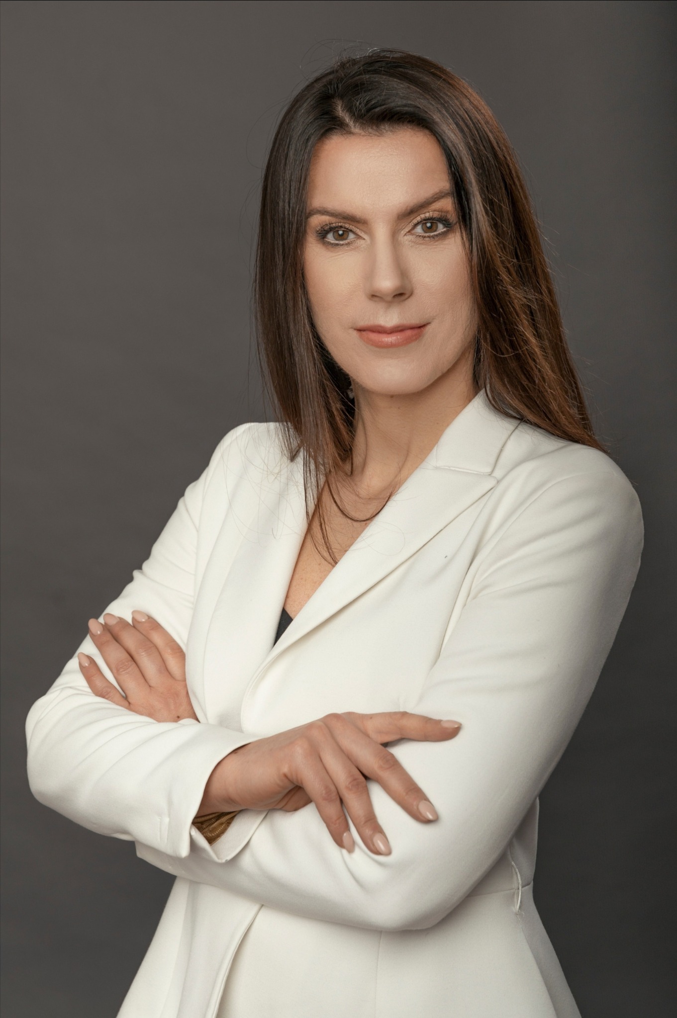 Portret kobiety z brązowymi włosami, ubranej w białą marynarkę, z założonymi rękami na szarym tle.
