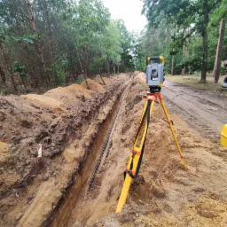 Pomiar geodezyjny wykopu w lesie z widocznymi kablami ułożonymi na dnie, teodolit na żółtym statywie ustawiony na skraju wykopu.