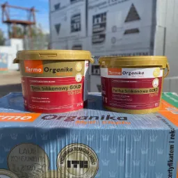 Dwa wiadra z tynkiem i farbą silikonową marki Termo Organika, linia GOLD, TO-TSG i TO-FSG, ustawione na opakowaniu zbiorczym z logo firmy, w tle widoczne płyty styropianowe i konstrukcja budowlana.