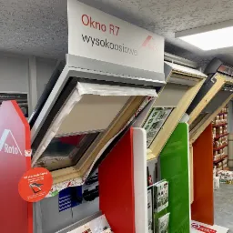 Ekspozycja okien dachowych Roto R7 z roletami wewnętrznymi w sklepie, widoczne materiały informacyjne i reklamy producenta, na półkach w tle widoczne materiały budowlane.