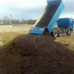 Ciężarówka z podniesioną skrzynią wyładowuje dużą pryzmę ciemnej ziemi na polu, widoczne drzewa w tle.