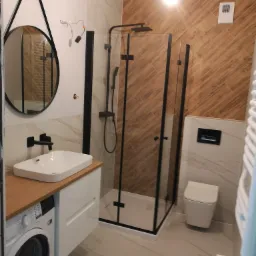Nowoczesna łazienka z kabiną prysznicową typu walk-in z czarnymi ramami, okrągłym lustrem z czarnym obramowaniem, białą umywalką nablatową na drewnianym blacie, pralką i wiszącą toaletą. Ściany...