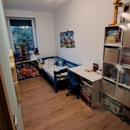 MasterDom - Pokój dziecięcy z drewnianą podłogą, niebieskim łóżkiem z motywem bajkowym, biurkiem i regałem z zabawkami, oświetlony lampą sufitową z kolorowymi żarówkami.