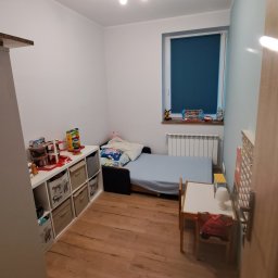 MasterDom - Pokój dziecięcy z drewnianą podłogą, białymi ścianami, niebieskim akcentem na jednej ścianie, łóżkiem z niebieską pościelą, białym regałem z zabawkami i drewnianym stolikiem z krzesłem, oświetlony...