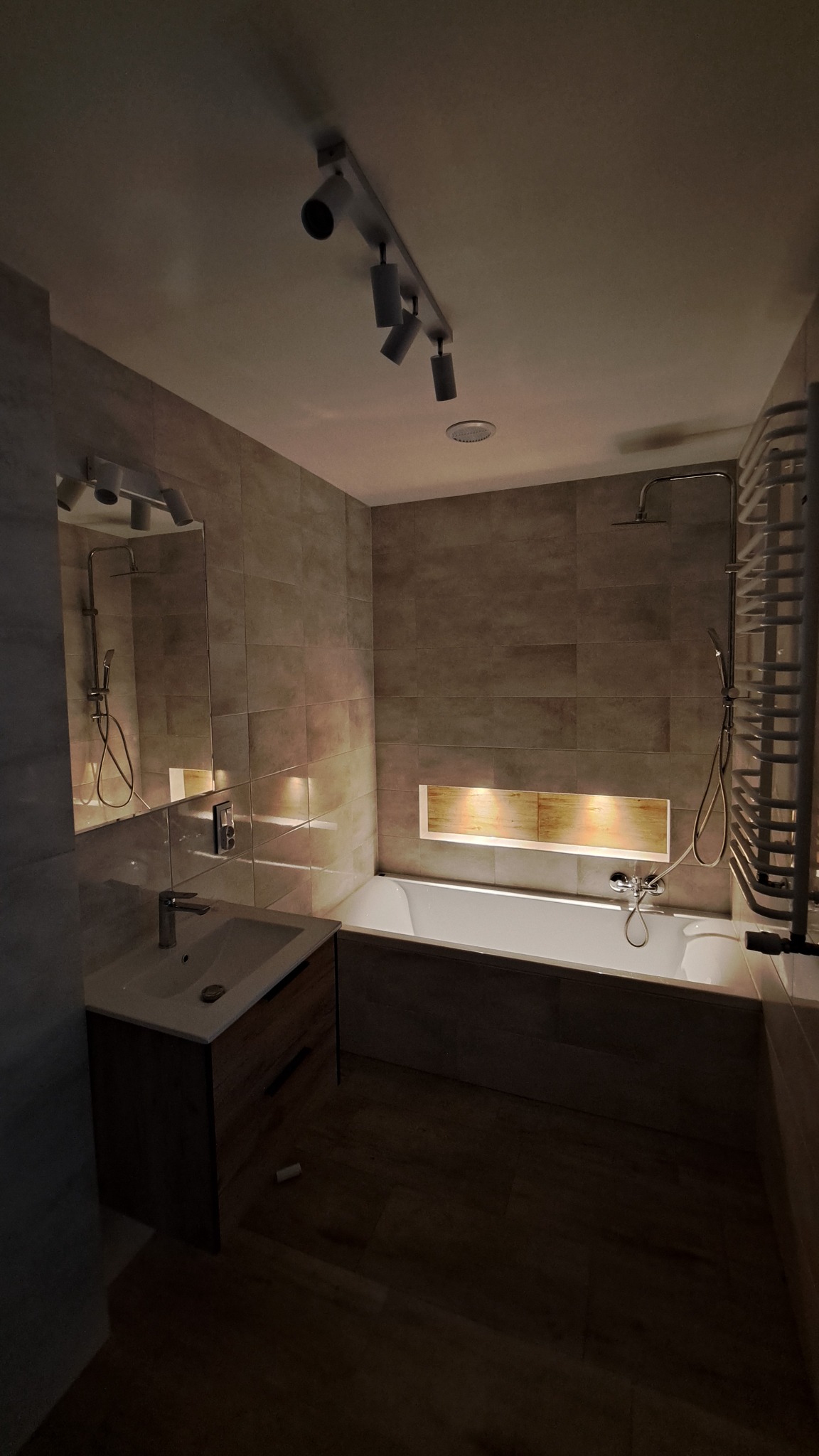 Nowoczesna łazienka z beżowymi płytkami, oświetleniem punktowym i wnęką nad wanną z ciepłym światłem, widok na umywalkę z szafką z drewnopodobnym frontem i grzejnik drabinkowy.