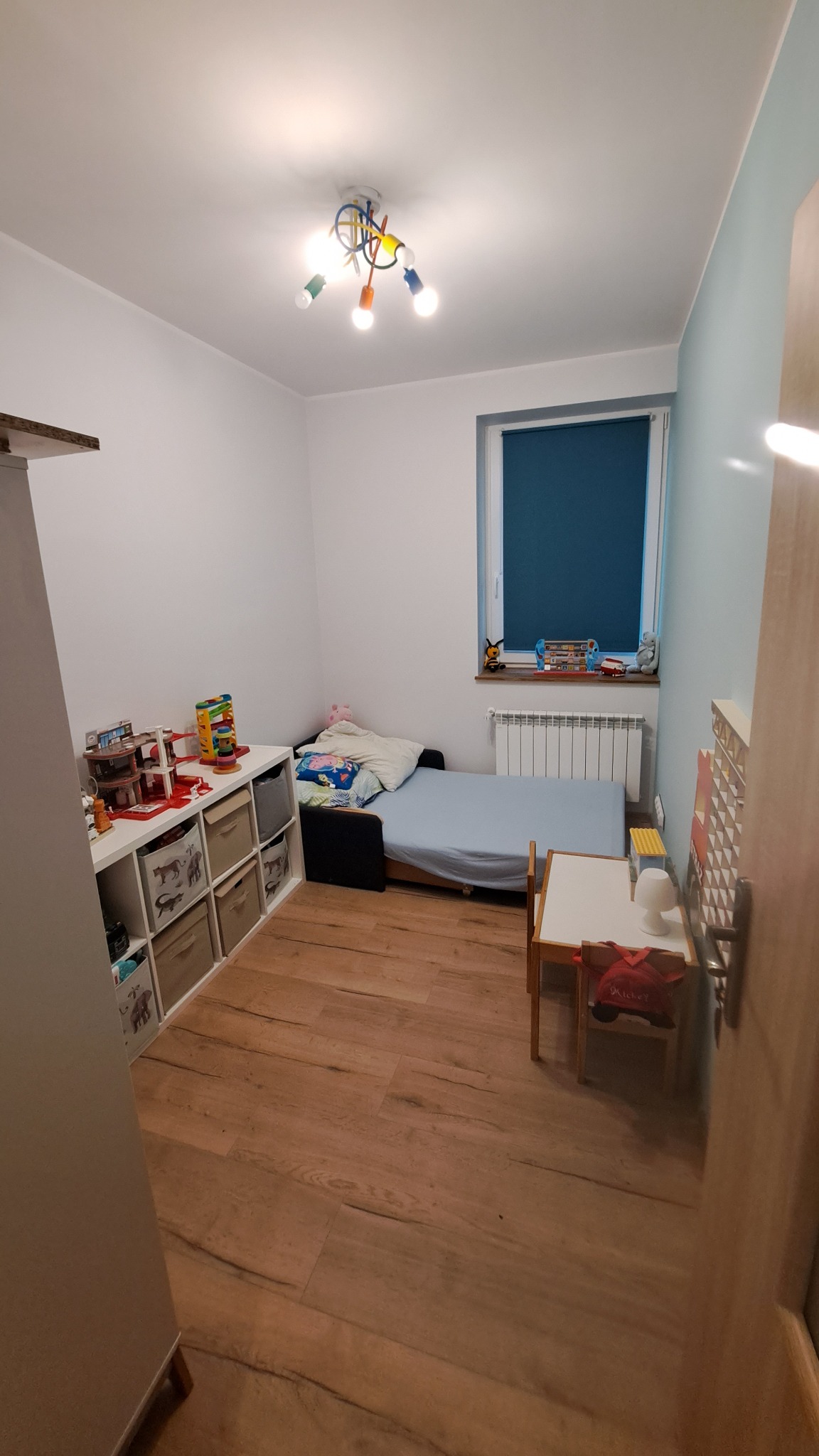 Pokój dziecięcy z drewnianą podłogą, białymi ścianami, niebieskim akcentem na jednej ścianie, łóżkiem z niebieską pościelą, białym regałem z zabawkami i drewnianym stolikiem z krzesłem, oświetlony...