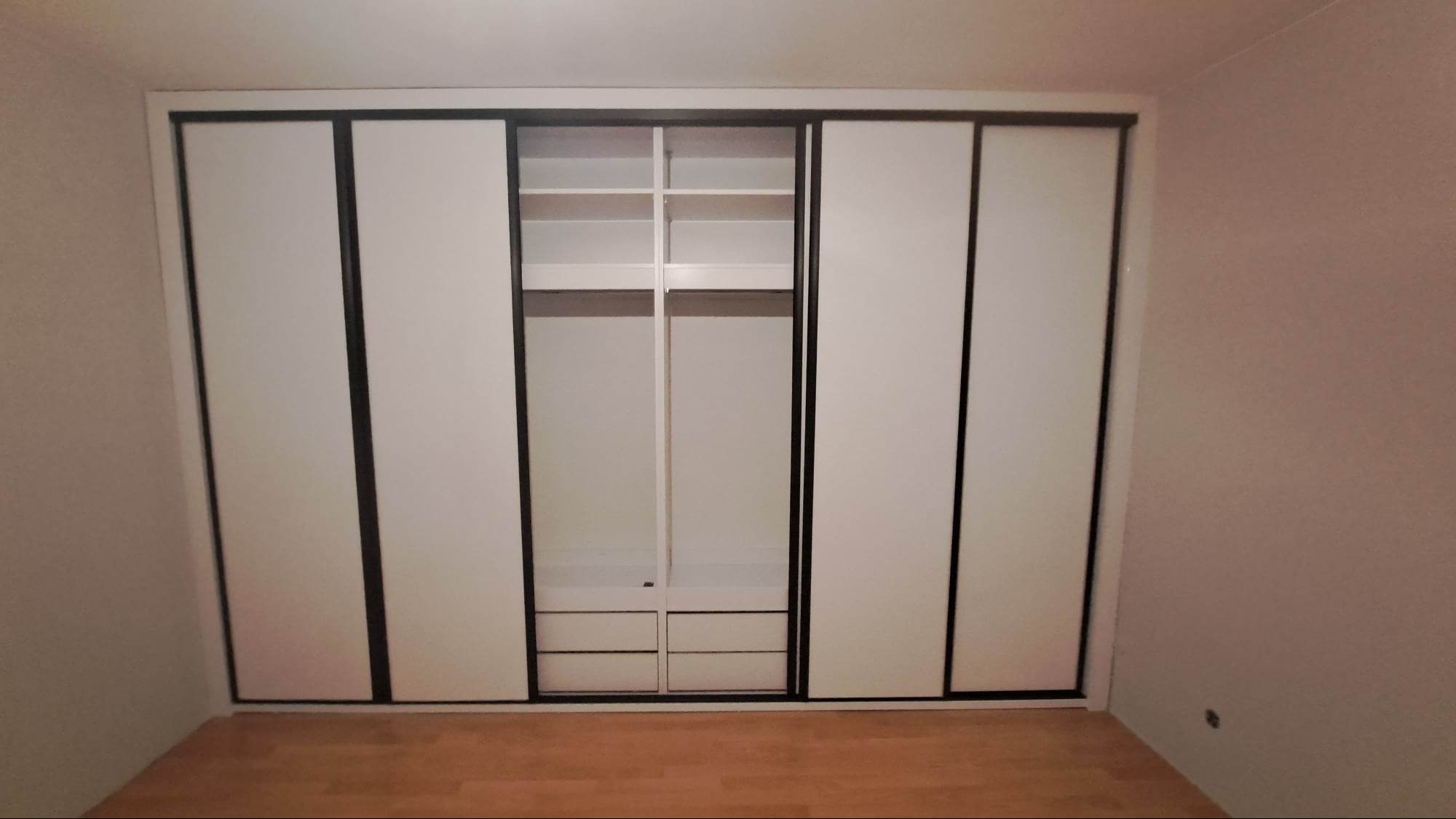 Wbudowana szafa z przesuwnymi białymi drzwiami i czarnymi ramami, częściowo otwarta, ukazująca półki i szuflady w środkowej części, na tle beżowej ściany i drewnianej podłogi.