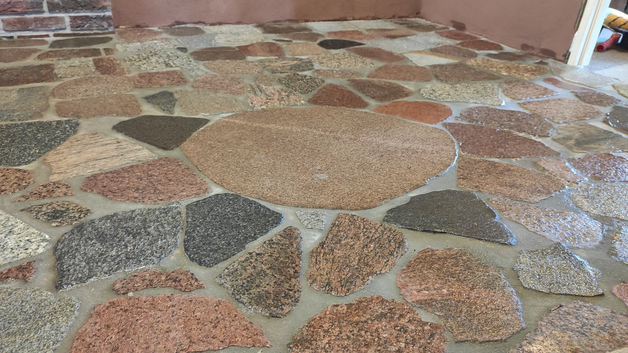 Kamienna podłoga z nieregularnych, wielobarwnych płyt, z centralnym, okrągłym elementem, na tle ceglanej ściany i fragmentu drzwi.