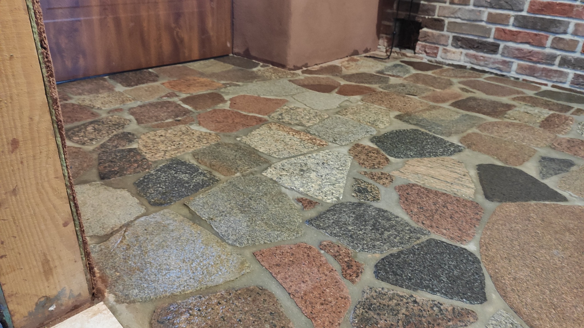 Kamienna posadzka z nieregularnych płytek w odcieniach szarości, brązu i czerwieni, ułożona wewnątrz pomieszczenia, widoczny fragment ściany z cegły i drewnianej ościeżnicy.
