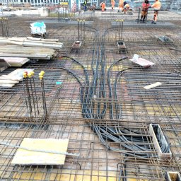 Tech.inBud - Budowa stropu: widok na zbrojenie z ułożonymi peszlami instalacji elektrycznej, w tle pracownicy budowlani w pomarańczowych kombinezonach.