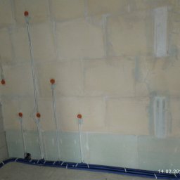 Tech.inBud - Ściana z surowego betonu komórkowego z widocznymi przewodami elektrycznymi w peszlach mocowanymi do ściany, przygotowanymi do podłączenia gniazdek i włączników, oraz niebieskimi rurami peszlowymi...