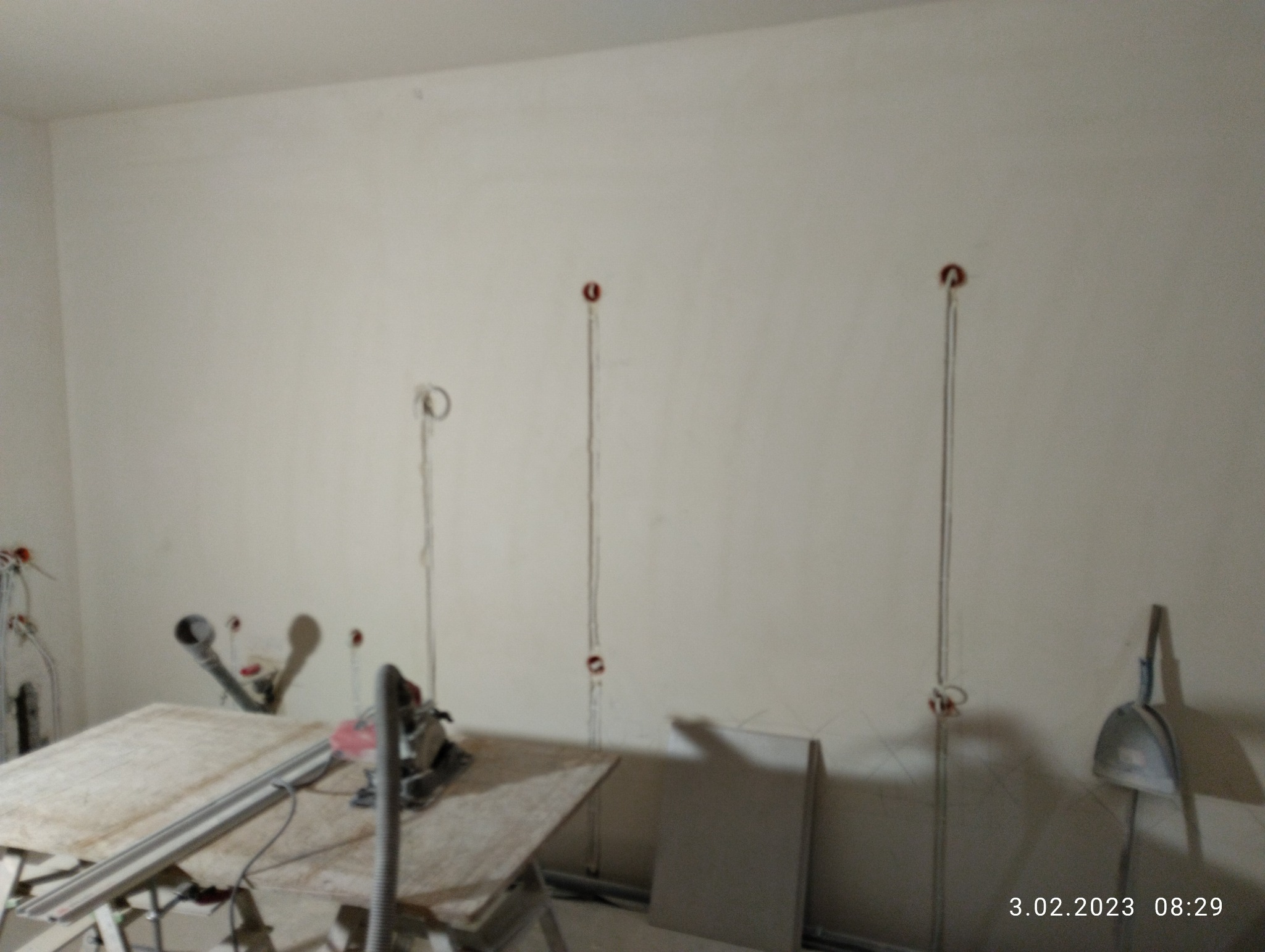 Wnętrze w trakcie prac instalacyjnych: widoczne kable elektryczne wystające ze ściany, rury, stół roboczy z piłą tarczową i odkurzaczem przemysłowym. Otwory na gniazdka elektryczne na różnych...