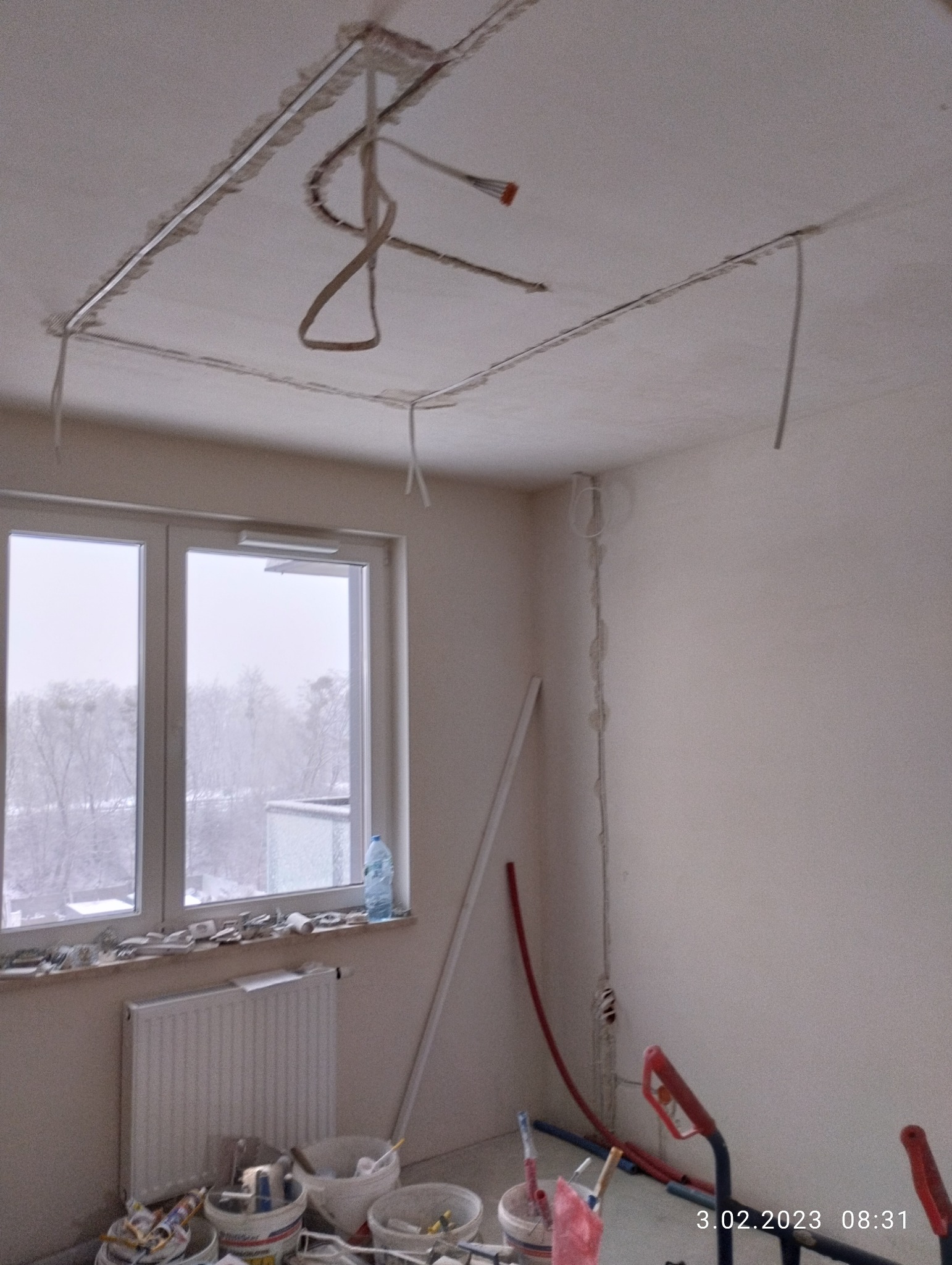 Pokój w trakcie remontu: przygotowane pod sufitem korytka na oświetlenie, wystające przewody, ślady po kuciu w ścianach, narzędzia i puszki z farbą przy oknie.
