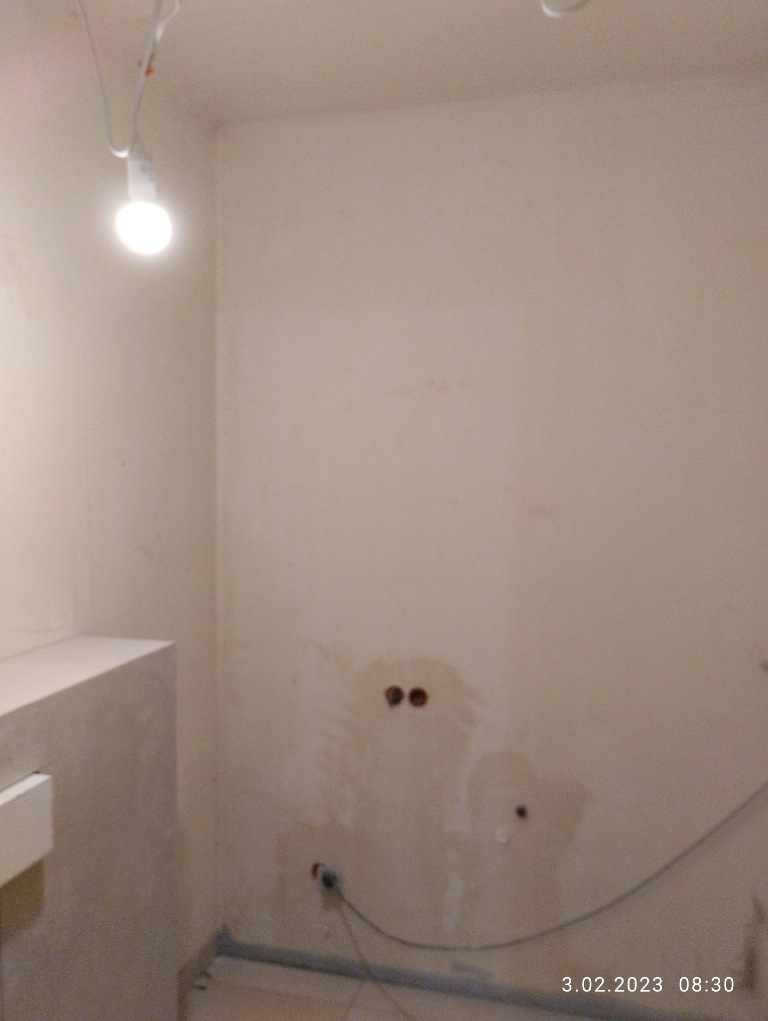 Ściana w trakcie przygotowania do malowania z widocznymi otworami po gniazdkach elektrycznych, podłączonym kablem i żarówką na kablu jako tymczasowe oświetlenie.