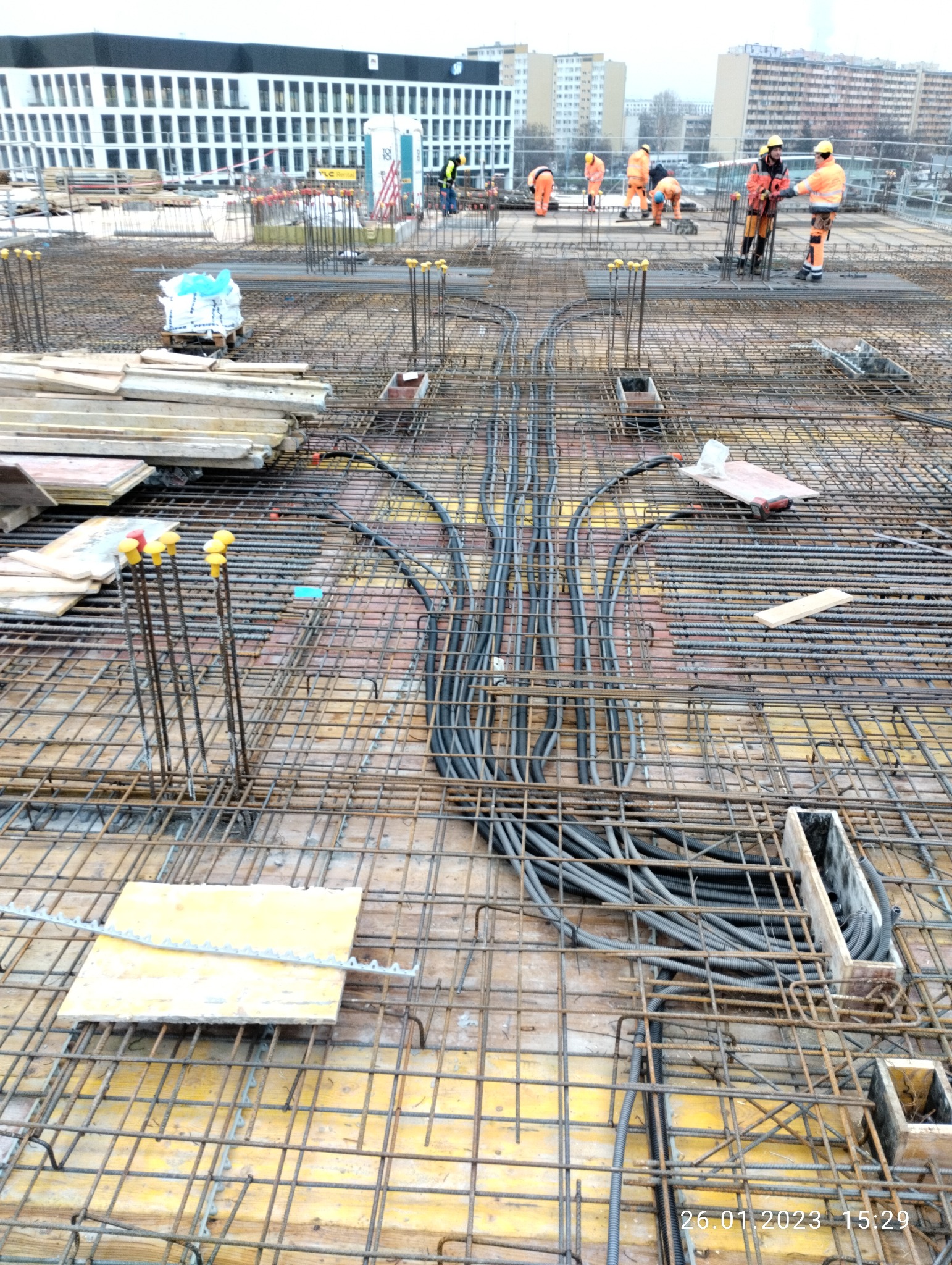 Budowa stropu: widok na zbrojenie z ułożonymi peszlami instalacji elektrycznej, w tle pracownicy budowlani w pomarańczowych kombinezonach.