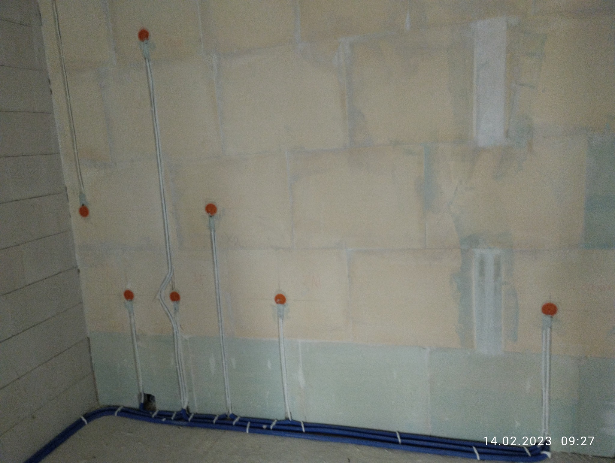 Ściana z surowego betonu komórkowego z widocznymi przewodami elektrycznymi w peszlach mocowanymi do ściany, przygotowanymi do podłączenia gniazdek i włączników, oraz niebieskimi rurami peszlowymi...