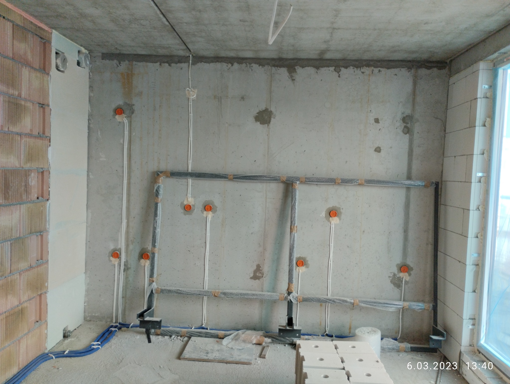 Surowy stan wnętrza z widocznymi przewodami elektrycznymi wychodzącymi ze ścian, zabezpieczonymi rurami instalacyjnymi i puszkami, nieotynkowana ściana z betonu komórkowego, fragment okna po prawej.