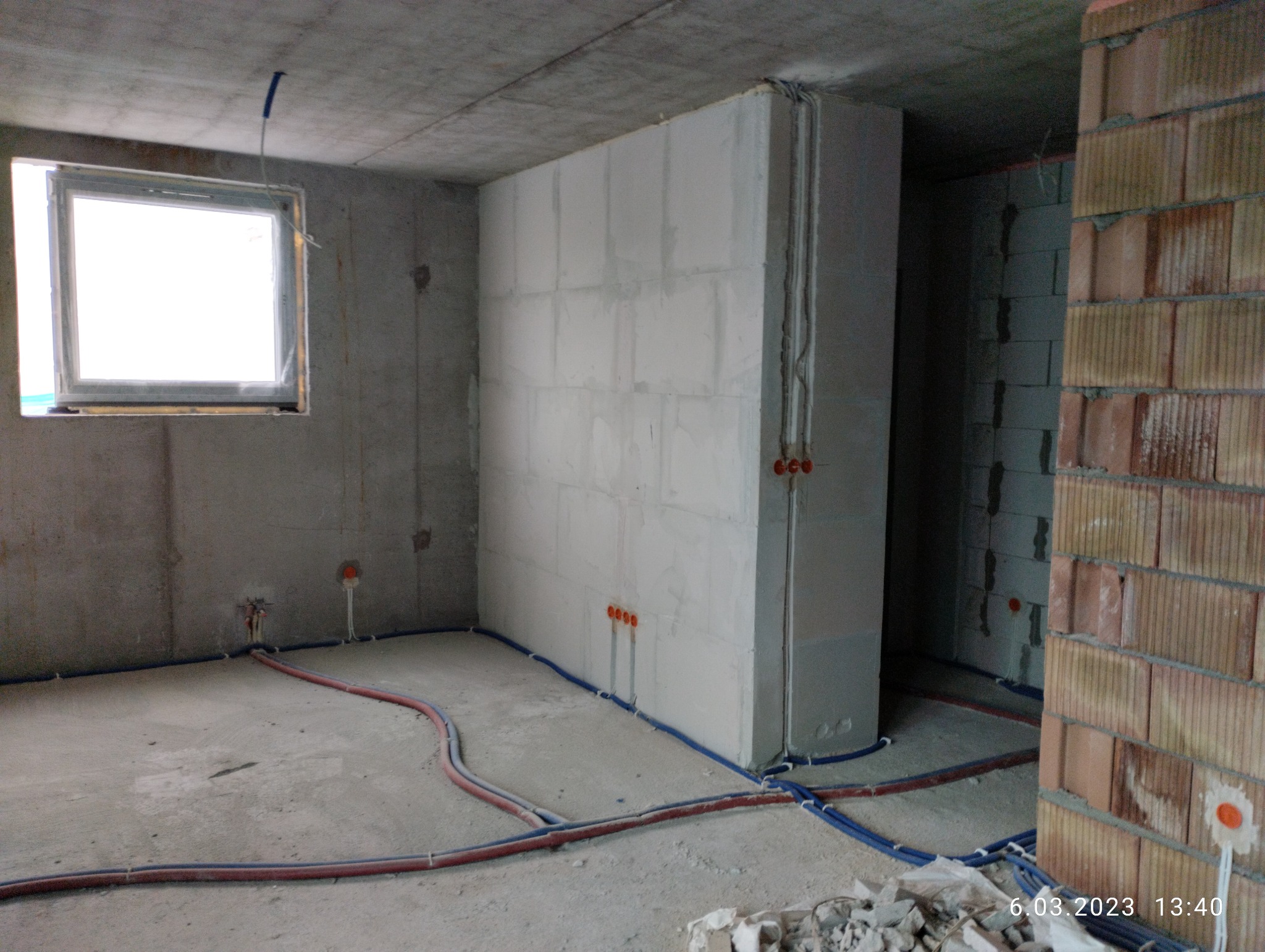 Surowy stan wnętrza w trakcie prac: widoczne betonowe ściany, okno, instalacje elektryczne w peszlach na podłodze i ścianach, oraz fragment ściany z czerwonej cegły.
