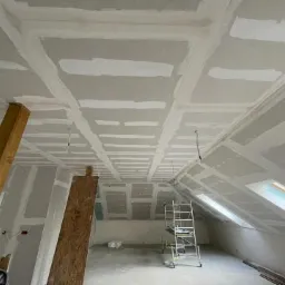 Poddasze w trakcie wykańczania, widoczne płyty gipsowo-kartonowe na skośnym suficie, zaszpachlowane łączenia, drewniane podpory i aluminiowa drabina, okno dachowe w tle.