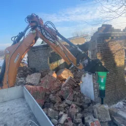 Pomarańczowa koparka CASE 695ST z hydraulicznym młotem wyburzeniowym SC-28, gruz ceglany i fragmenty muru po rozbiórce budynku w zimowej scenerii.