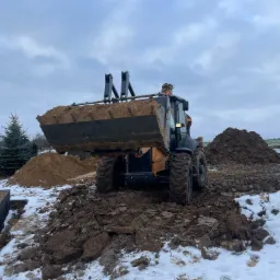 Żółto-czarna koparko-ładowarka z pełną łyżką ziemi na budowie w zimowej scenerii, widoczny fragment fundamentu i pracownik w oddali.