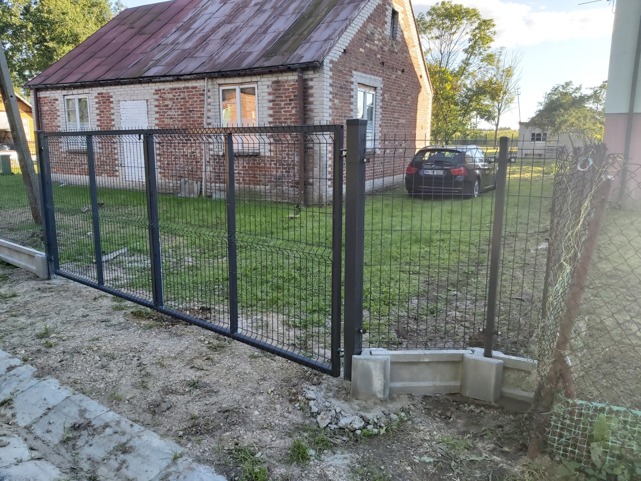 Nowo zainstalowana brama z paneli ogrodzeniowych na posesji z ceglanym domem i samochodem w tle, widoczny fragment starego ogrodzenia z siatki.