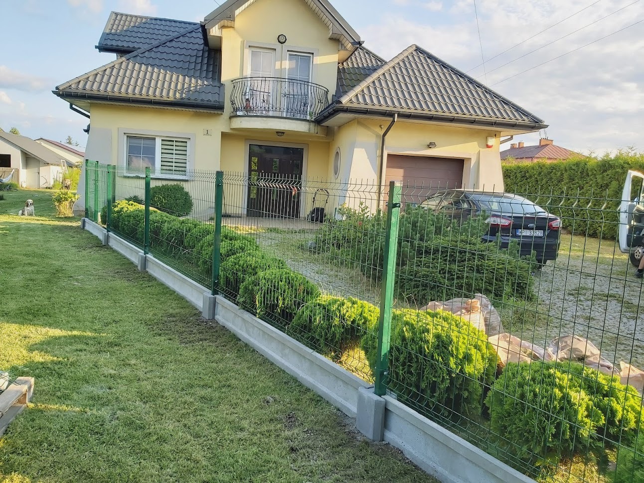 Zielone ogrodzenie panelowe z podmurówką betonową, otaczające posesję z żółtym domem i zadbanym trawnikiem, widok z zewnątrz.