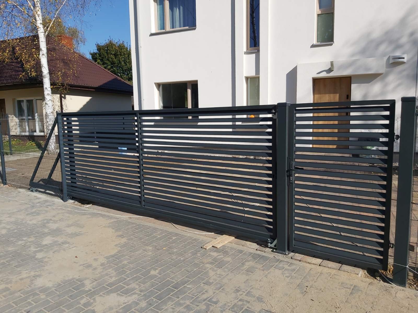 Nowoczesna, przesuwna brama wjazdowa z poziomymi panelami, pomalowana na grafitowy kolor, z widocznym ogrodzeniem z siatki w tle i fragmentem białego domu z drewnianymi drzwiami wejściowymi.