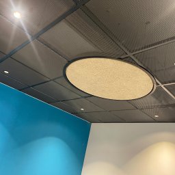 Fast Build by Roman Vynnyk - Metalowa siatka sufitowa z okrągłym panelem akustycznym w kolorze beżowym, oświetlenie punktowe i fragmenty błękitnej i białej ściany.