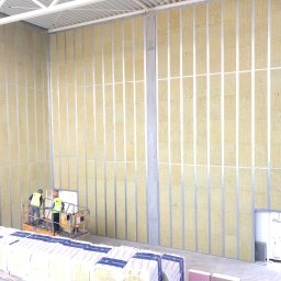 Fast Build by Roman Vynnyk - Montaż izolacji akustycznej w metalowej konstrukcji szkieletowej wewnątrz budynku, widoczni dwaj pracownicy na podnośniku, stos materiałów izolacyjnych na pierwszym planie.