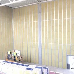 Montaż izolacji akustycznej w metalowej konstrukcji szkieletowej wewnątrz budynku, widoczni dwaj pracownicy na podnośniku, stos materiałów izolacyjnych na pierwszym planie.