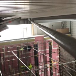 Fast Build by Roman Vynnyk - Montaż rusztowania wewnątrz pomieszczenia, widoczne płyty izolacyjne na ścianach i metalowa konstrukcja sufitu, pracownicy w odblaskowych ubraniach.