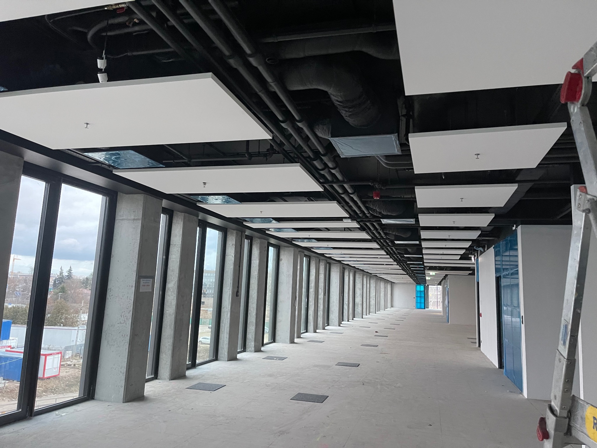 Długi korytarz biurowy w trakcie remontu z widocznymi sufitami podwieszanymi kasetonowymi, instalacjami pod sufitem i betonowymi słupami przy oknach.