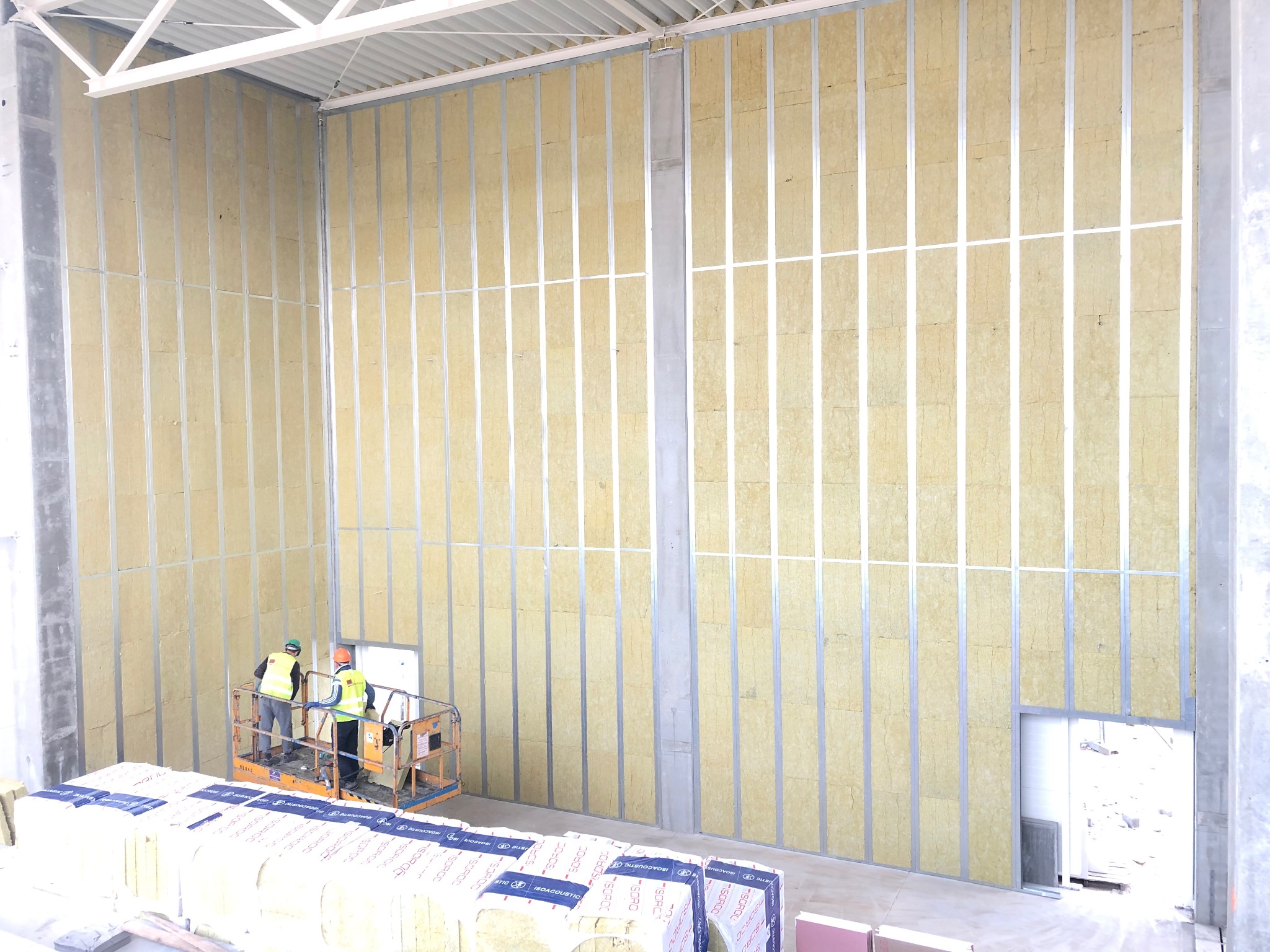 Montaż izolacji akustycznej w metalowej konstrukcji szkieletowej wewnątrz budynku, widoczni dwaj pracownicy na podnośniku, stos materiałów izolacyjnych na pierwszym planie.