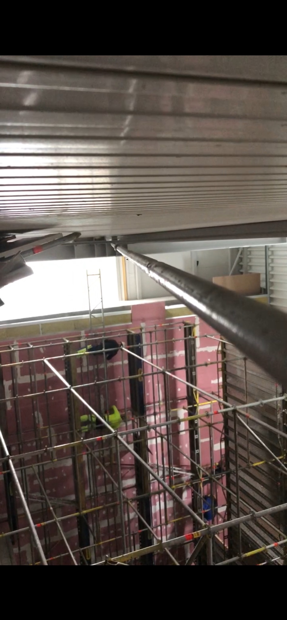 Montaż rusztowania wewnątrz pomieszczenia, widoczne płyty izolacyjne na ścianach i metalowa konstrukcja sufitu, pracownicy w odblaskowych ubraniach.