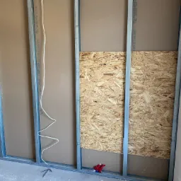 Montaż ściany działowej: widoczne metalowe profile, płyta OSB i częściowo zamontowana płyta gipsowo-kartonowa z widocznym nadrukiem producenta, instalacja elektryczna w puszce.