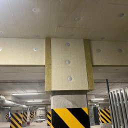 SAVRAN Sp.z o.o. - Ocieplenie poddasza w garażu podziemnym, widoczne płyty izolacyjne z wełny mineralnej przymocowane do sufitu i belek betonowych za pomocą okrągłych łączników, słupy z czarno-żółtymi pasami...